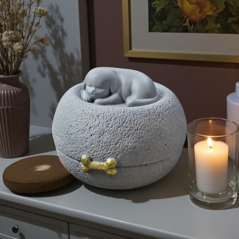 Grijze honden urn: een vredig slapend hondje bovenop een ronde, steenachtige basis, met een subtiel gouden botje als detail.